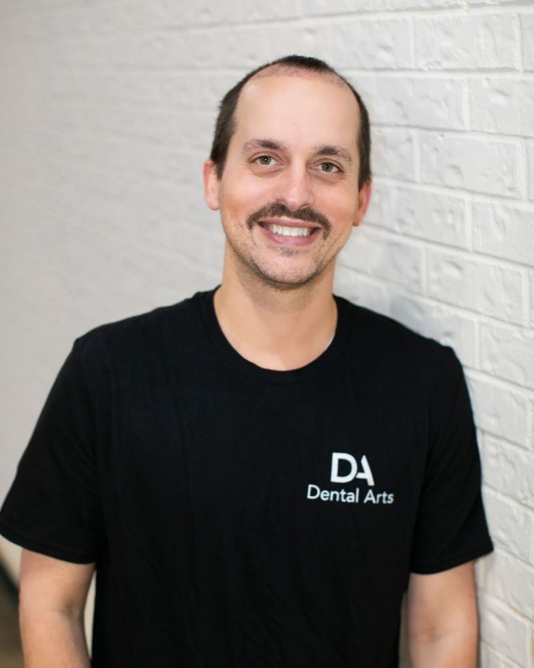 A man wearing glasses and a black shirt is standing in front of a white brick wall.