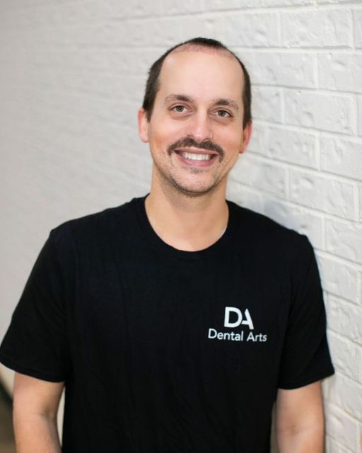 A man with a mustache is wearing a black dental arts shirt
