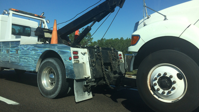 Tow truck towing a white semi-truck on the side of a road. Orange cone present.