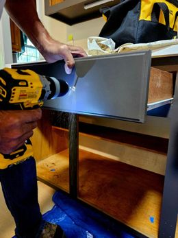 A man is using a drill to fix a drawer in a kitchen.