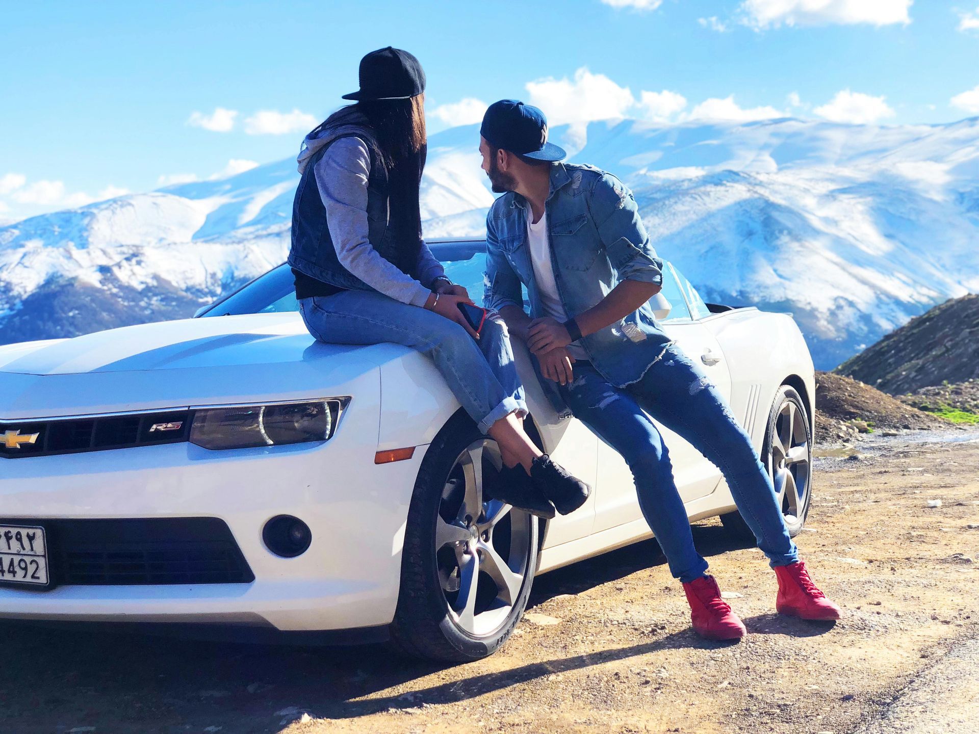 Two people in casual attire pose on a white sports car in a snowy mountain landscape.