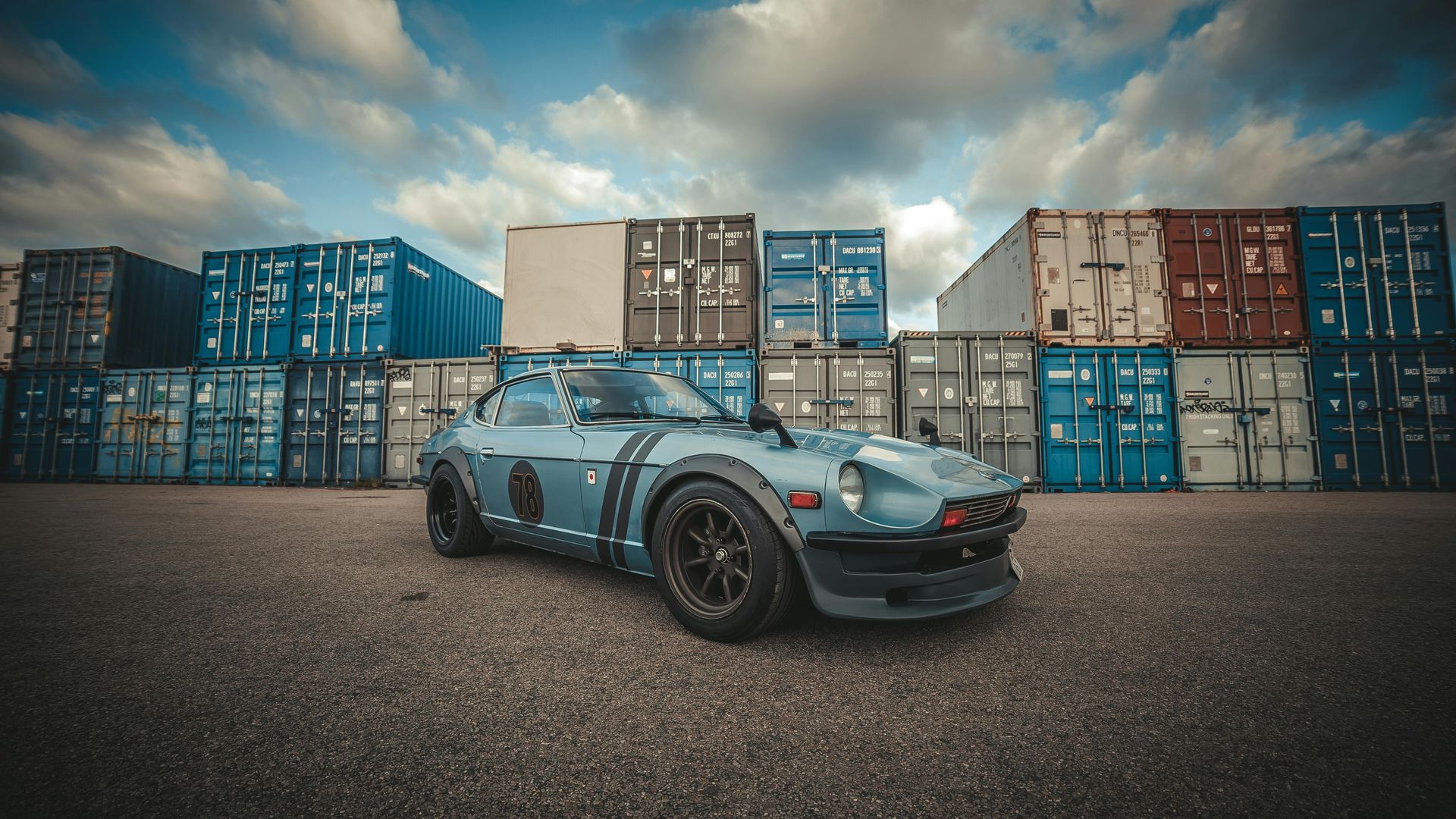Custom blue Datsun sports car parked in front of stacked shipping containers under a cloudy sky.