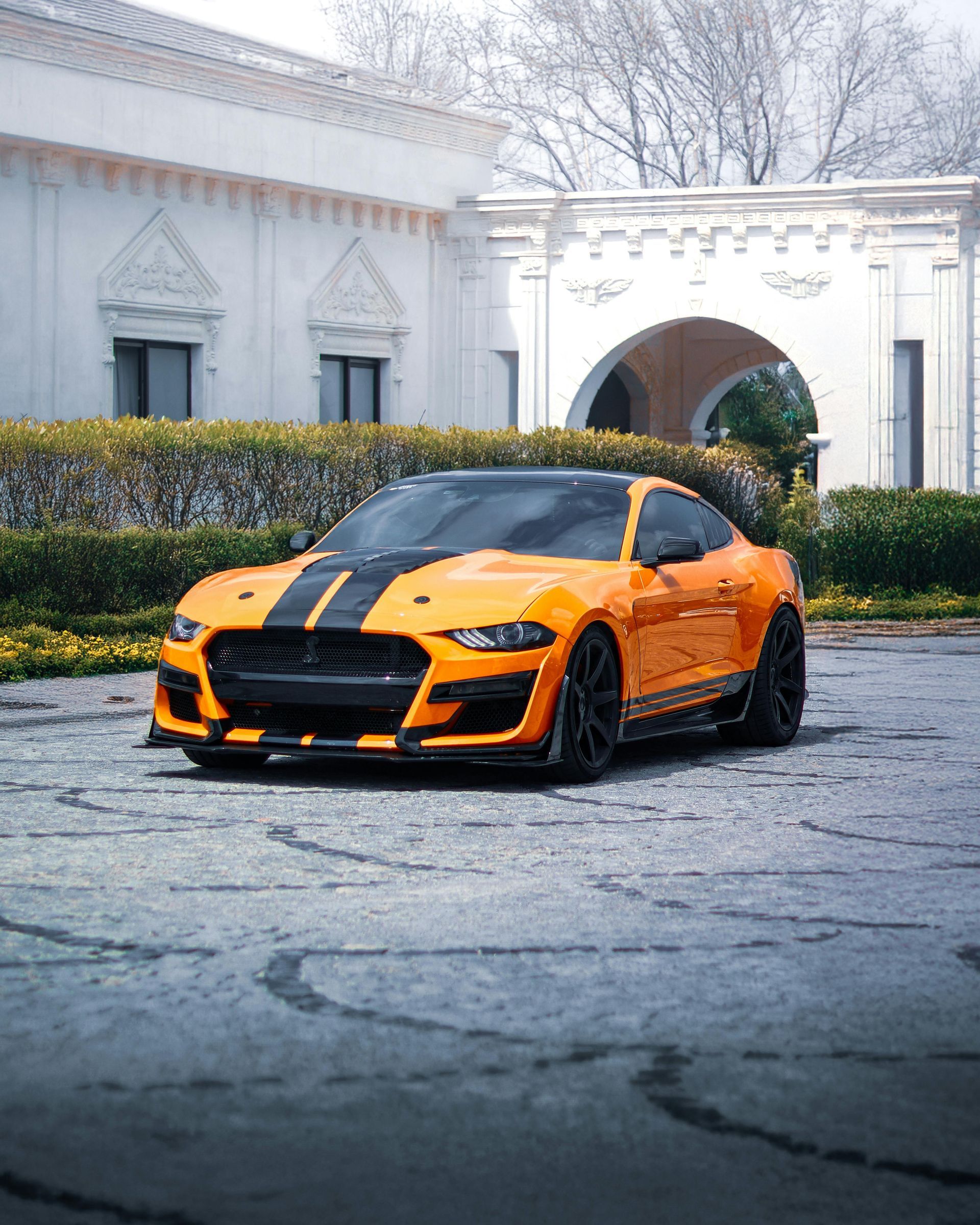 Orange Ford Mustang with black racing stripes parked on cobblestones in front of a white building.