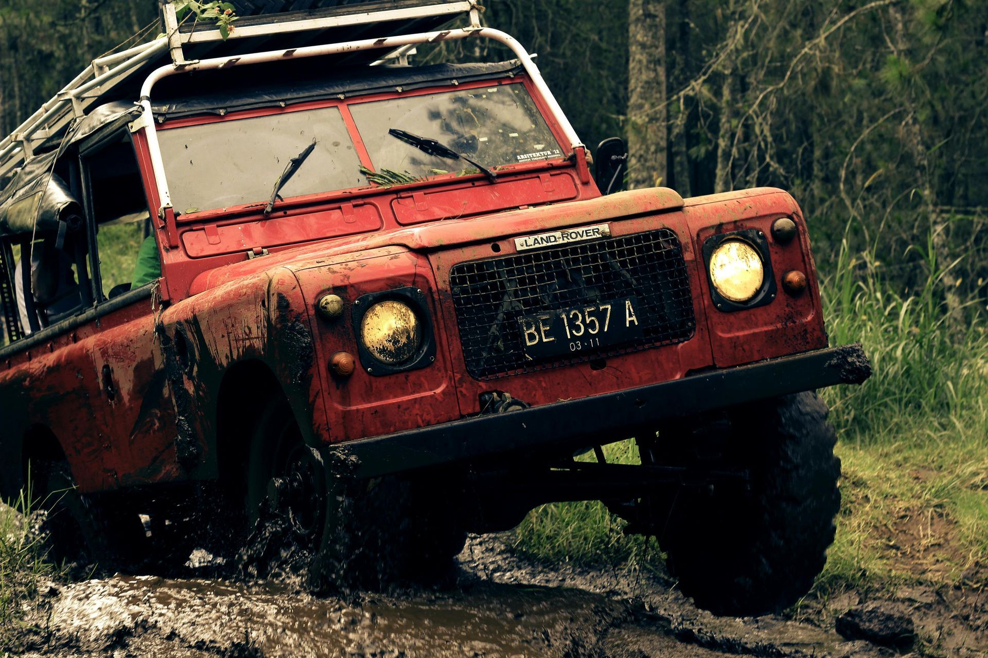 Red Land Rover off-roading through muddy terrain in a forest, splashing mud.