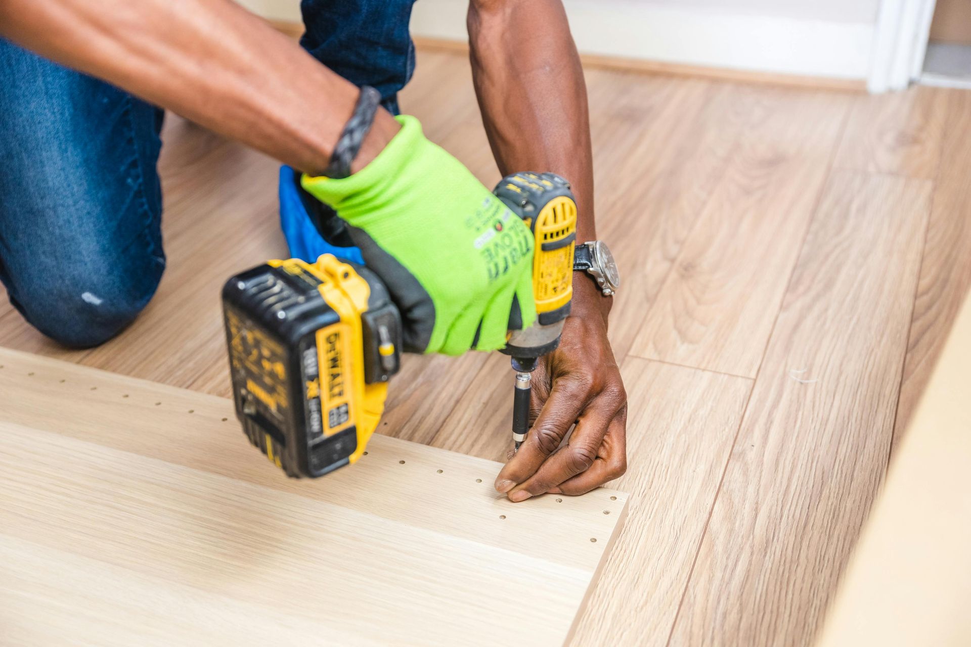 Person uses a power drill to install light-colored wood flooring, wearing a green glove.