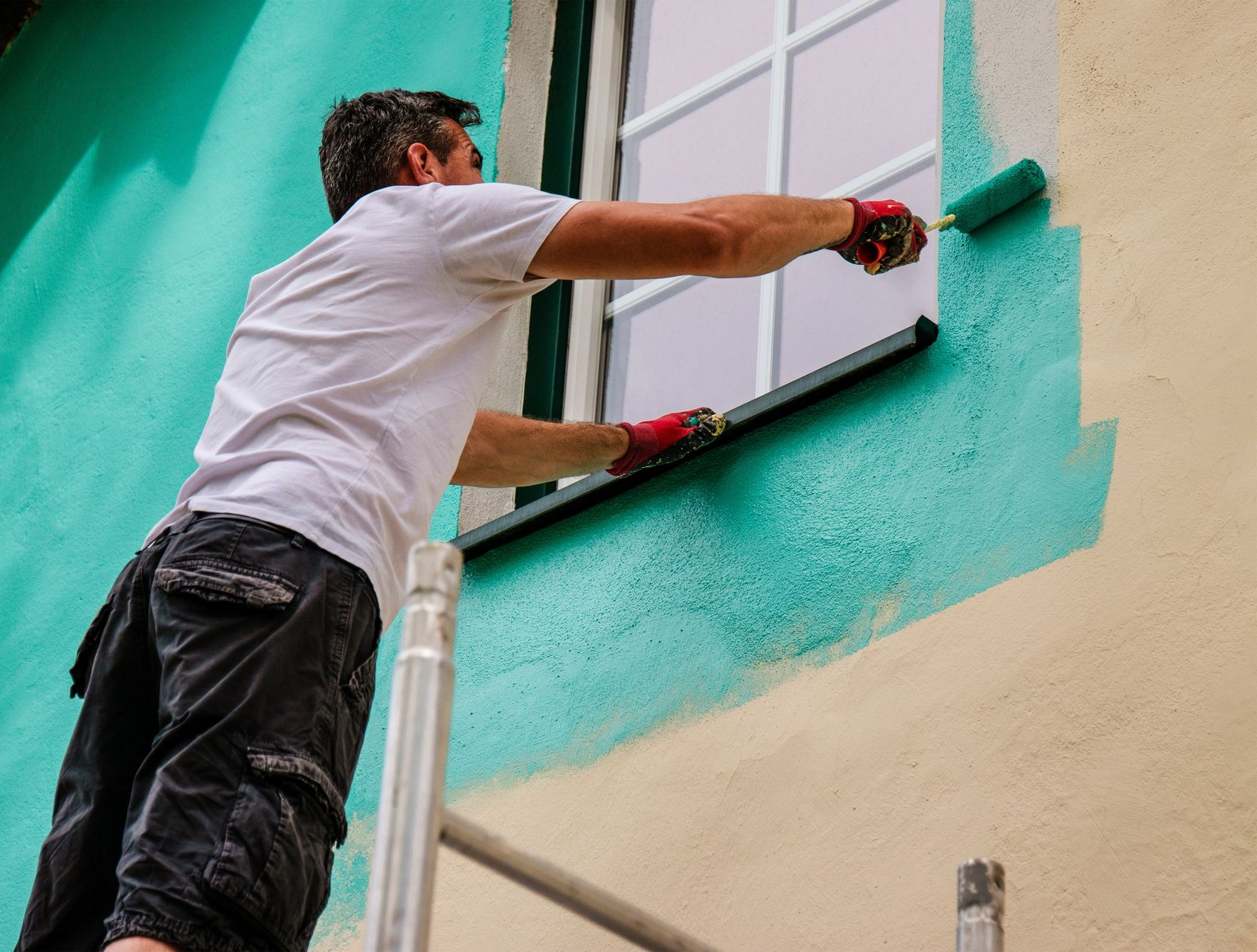 Man painting exterior wall turquoise, standing on scaffolding near window.