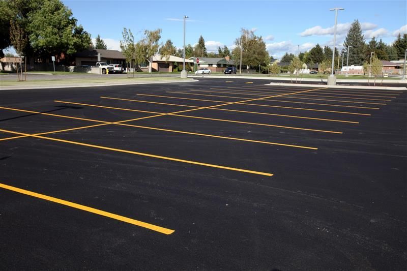 Empty asphalt parking lot with yellow painted parking space lines. Houses and trees in background.