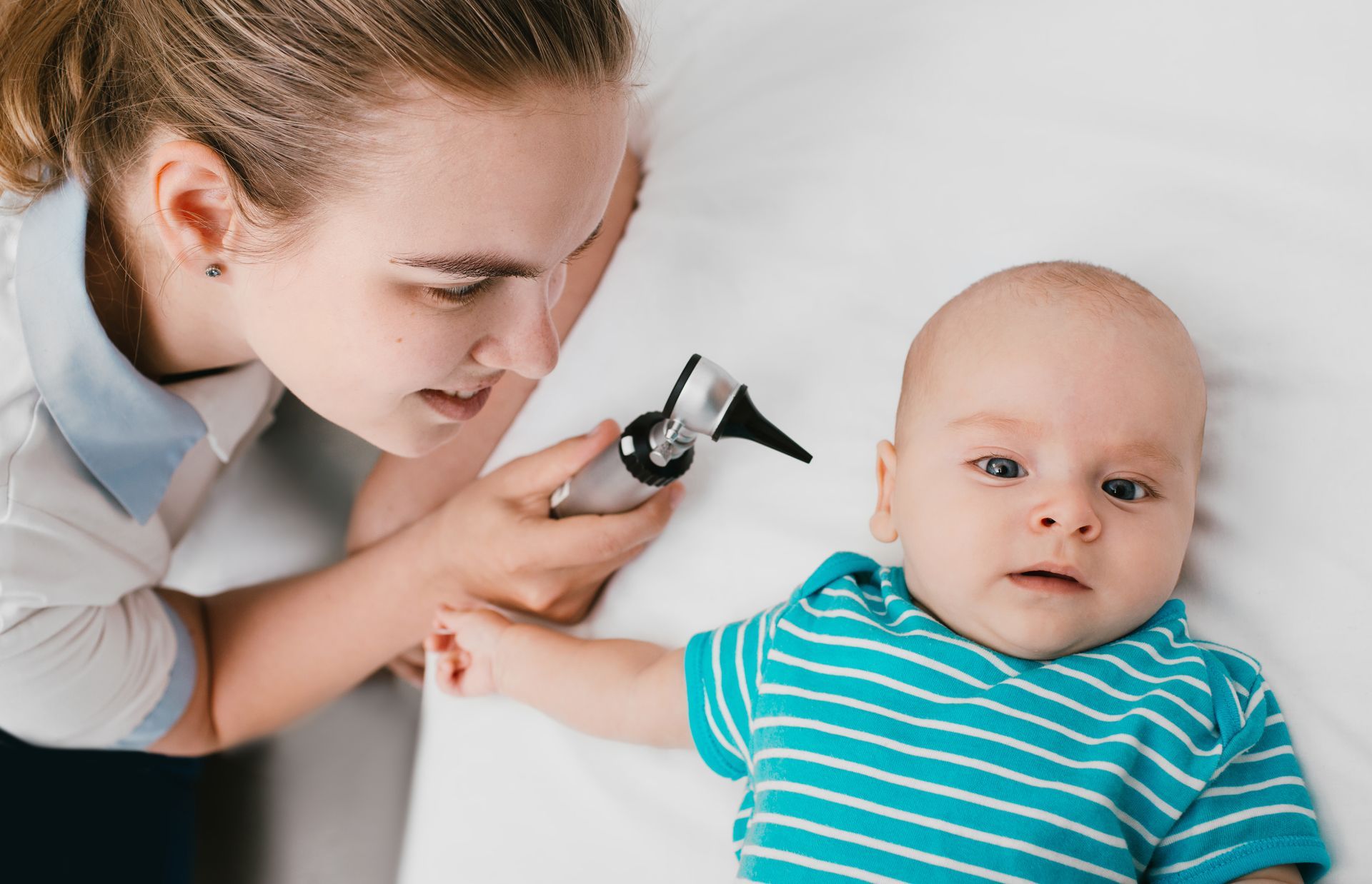 A female pediatrician is using an otoscope to look into a baby's ear. The baby is lying on a medical table with a bright, striped shirt on.