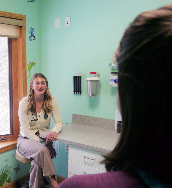 A woman with a stethoscope is sitting at a desk talking to another woman.