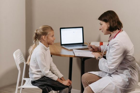 A young girl sits across a table from a doctor with a stethoscope. The doctor is taking notes near an open laptop.