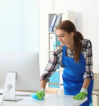 A woman is cleaning a desk in front of a computer in an office.