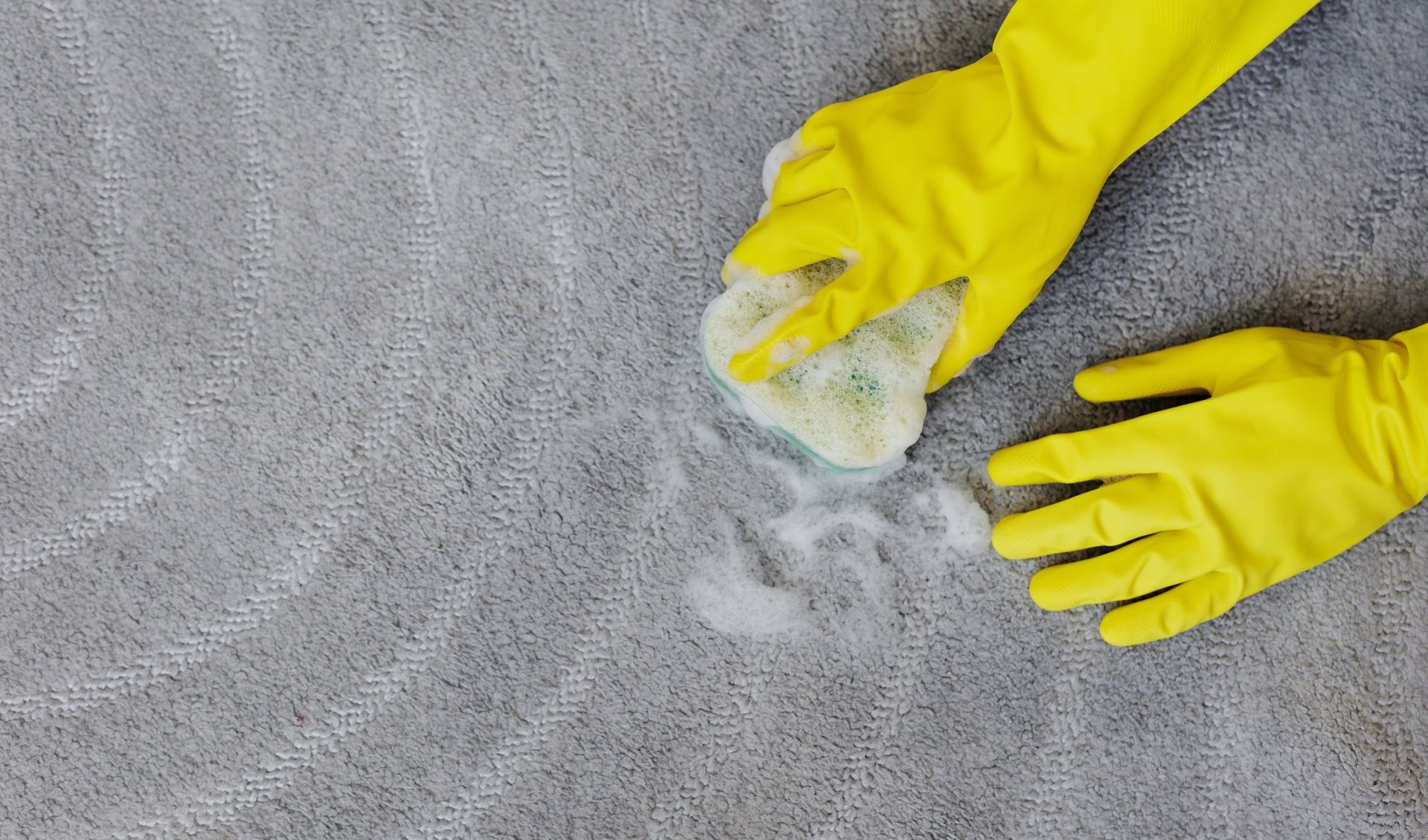 A person wearing yellow rubber gloves is cleaning a carpet with a cloth.