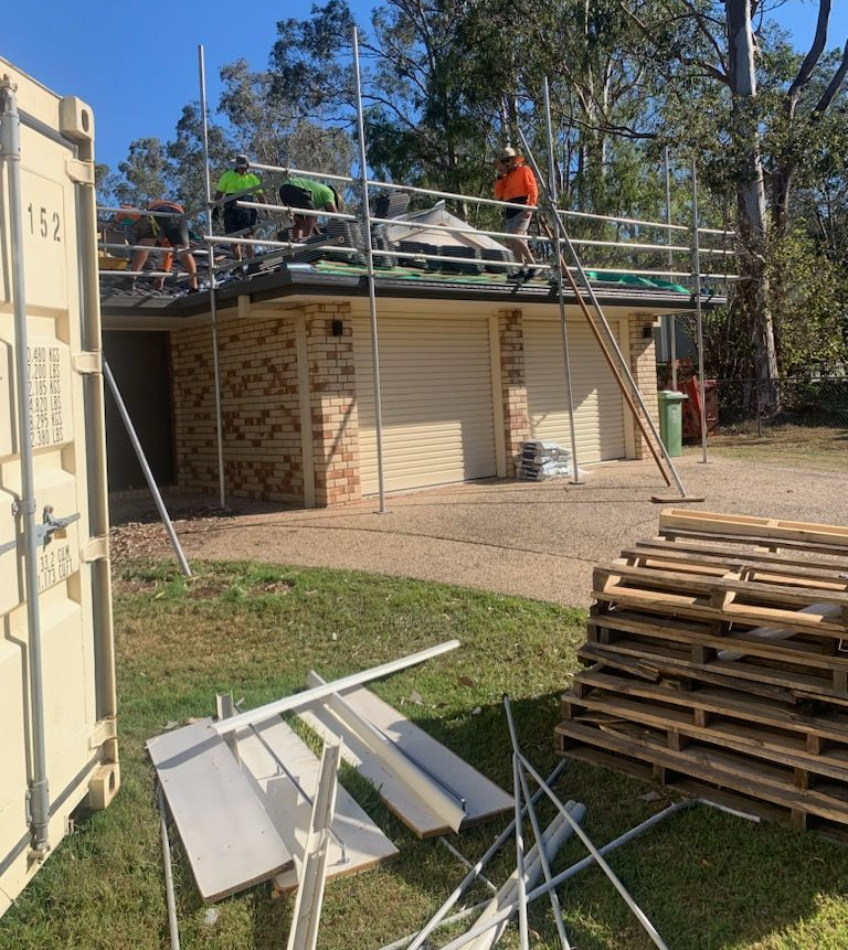 Construction workers on scaffolding repairing a brick building's roof; materials and pallets nearby. Sunny outdoor setting. — Boostn Pty Ltd in Glastonbury, QLD