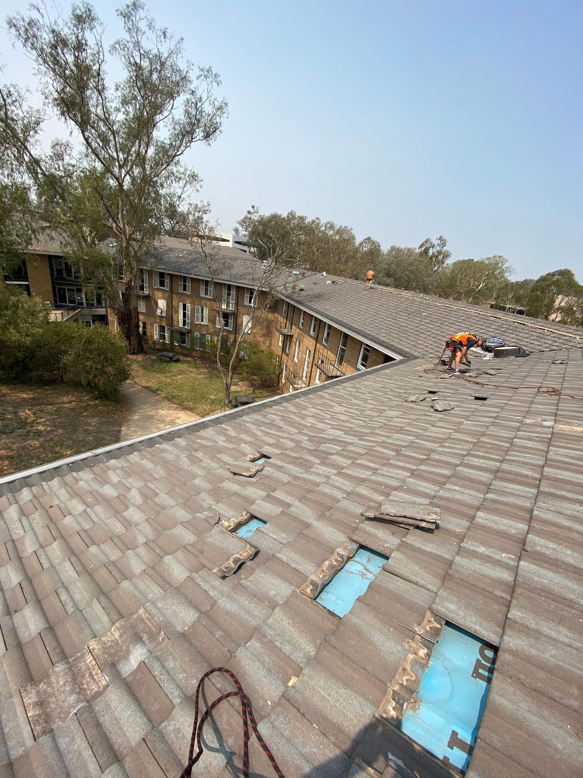Roofer repairing a damaged, shingled roof with exposed blue underlayment, next to an apartment building, on a sunny day. — Boostn Pty Ltd in Glastonbury, QLD