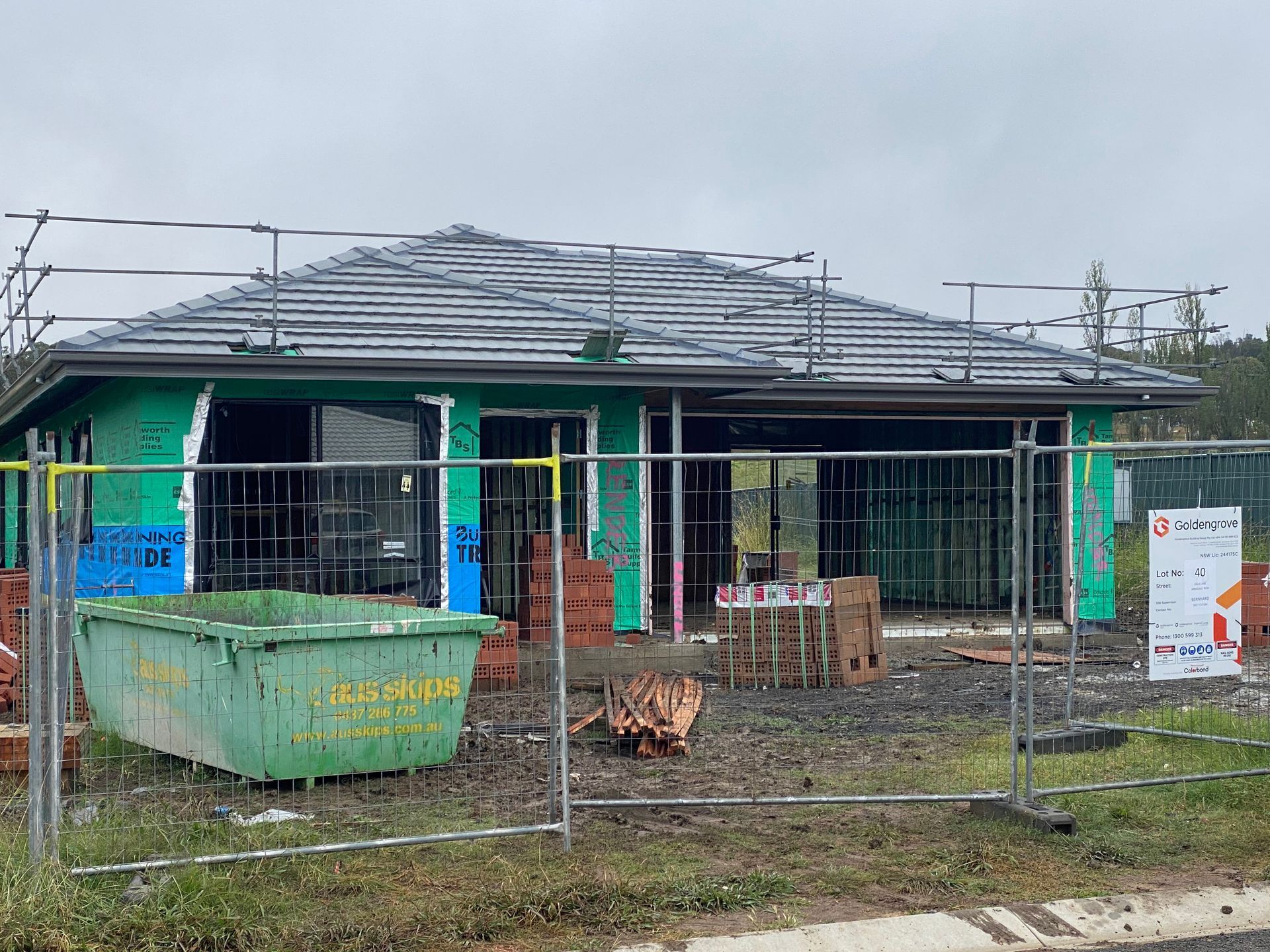 House under construction; green siding, gray roof with scaffolding. Green dumpster, brick, and metal fence in front. — Boostn Pty Ltd in Glastonbury, QLD