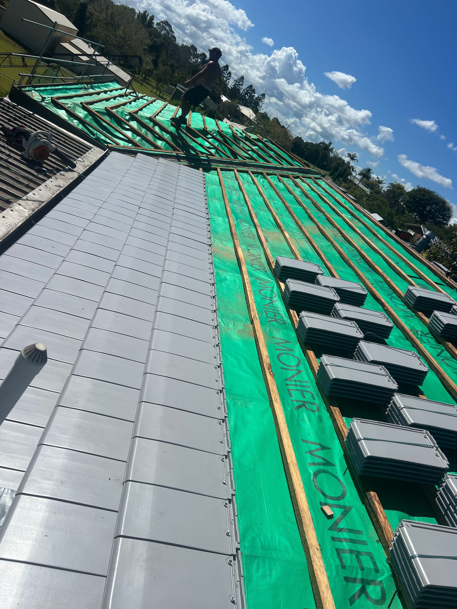 Roofers installing gray tiles on a roof. Green underlayment and bundles of tiles are visible. Blue sky in the background. — Boostn Pty Ltd in Glastonbury, QLD