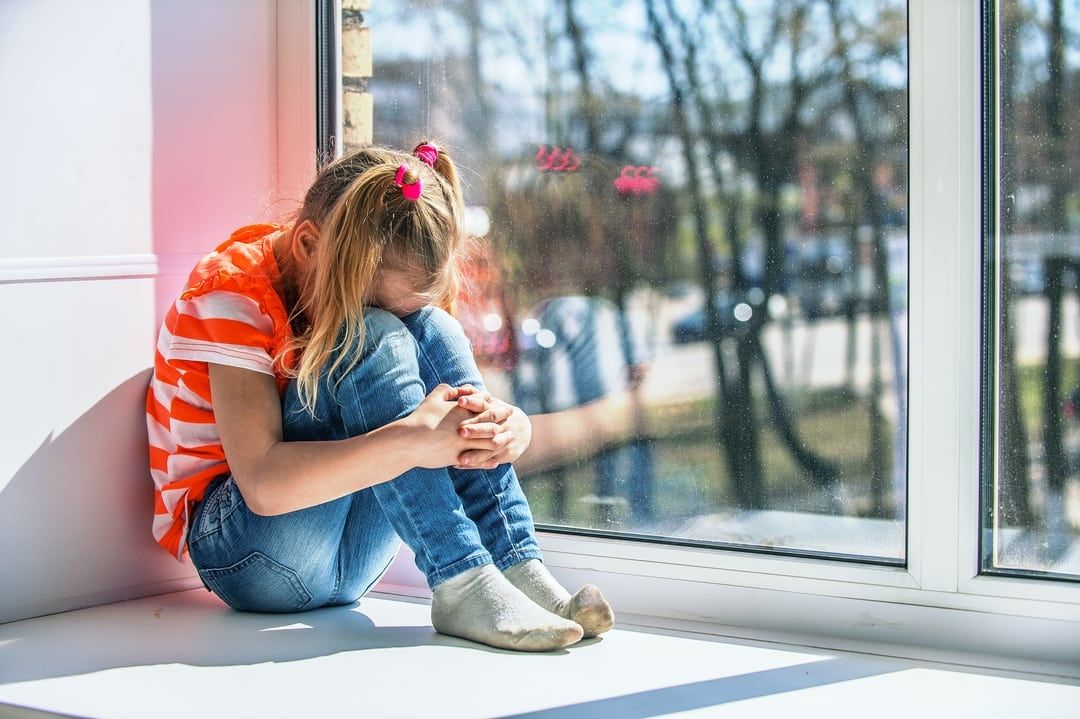 a little girl sits on a window sill with her head down