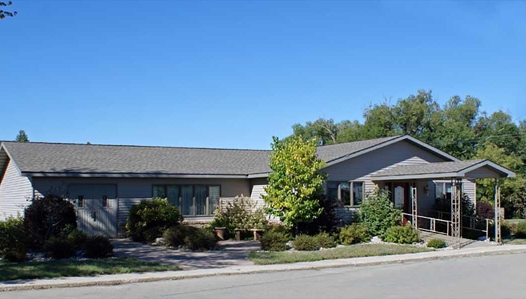 a house with a gray roof is surrounded by trees and bushes on a sunny day .