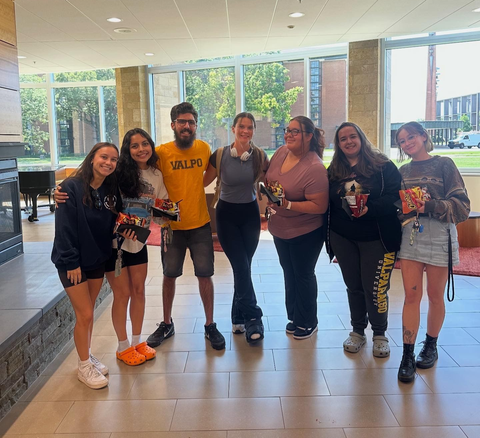 Seven people pose together in a bright, sunlit room with large windows, smiling and holding small snack bags.