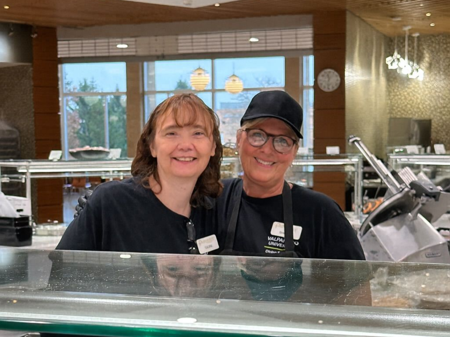 Two people smiling behind a counter in a food service area. One wears a black cap and glasses.