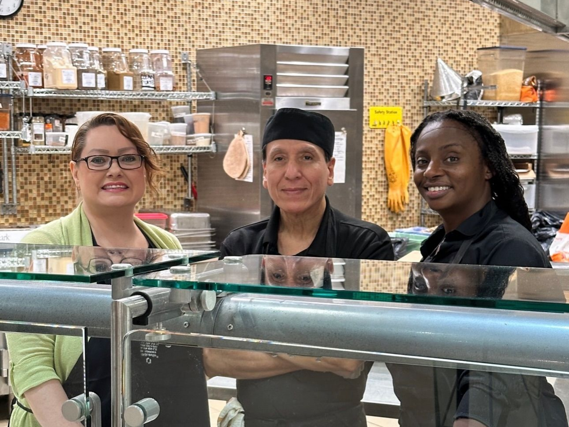 Three people behind a food counter: one with glasses, one in a chef's hat, and one smiling.