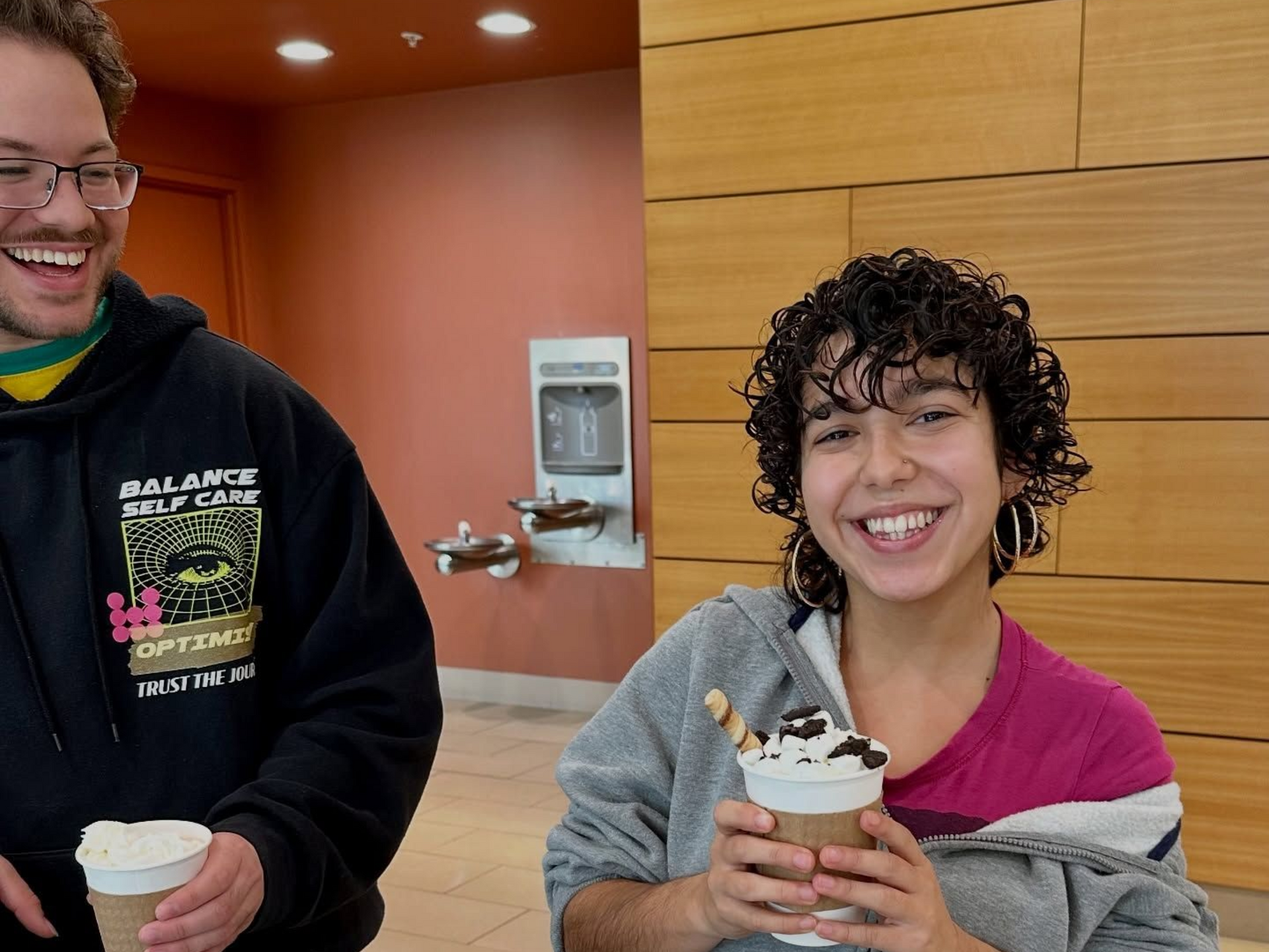 Two people smiling, holding coffee cups. The woman has curly hair and a decorative drink.
