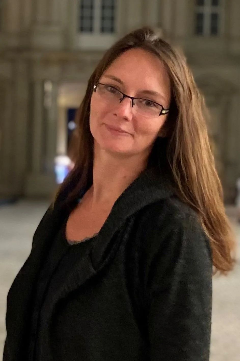 Eine Frau mit Brille und schwarzer Jacke steht vor einem Gebäude.