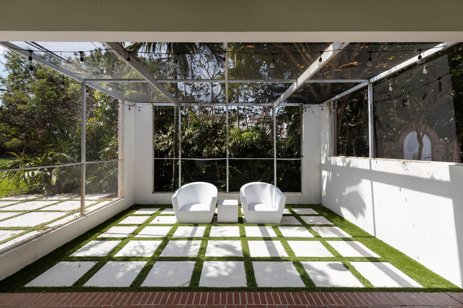 Outdoor seating area with white chairs and table on a square-patterned lawn, enclosed by glass walls and roof.