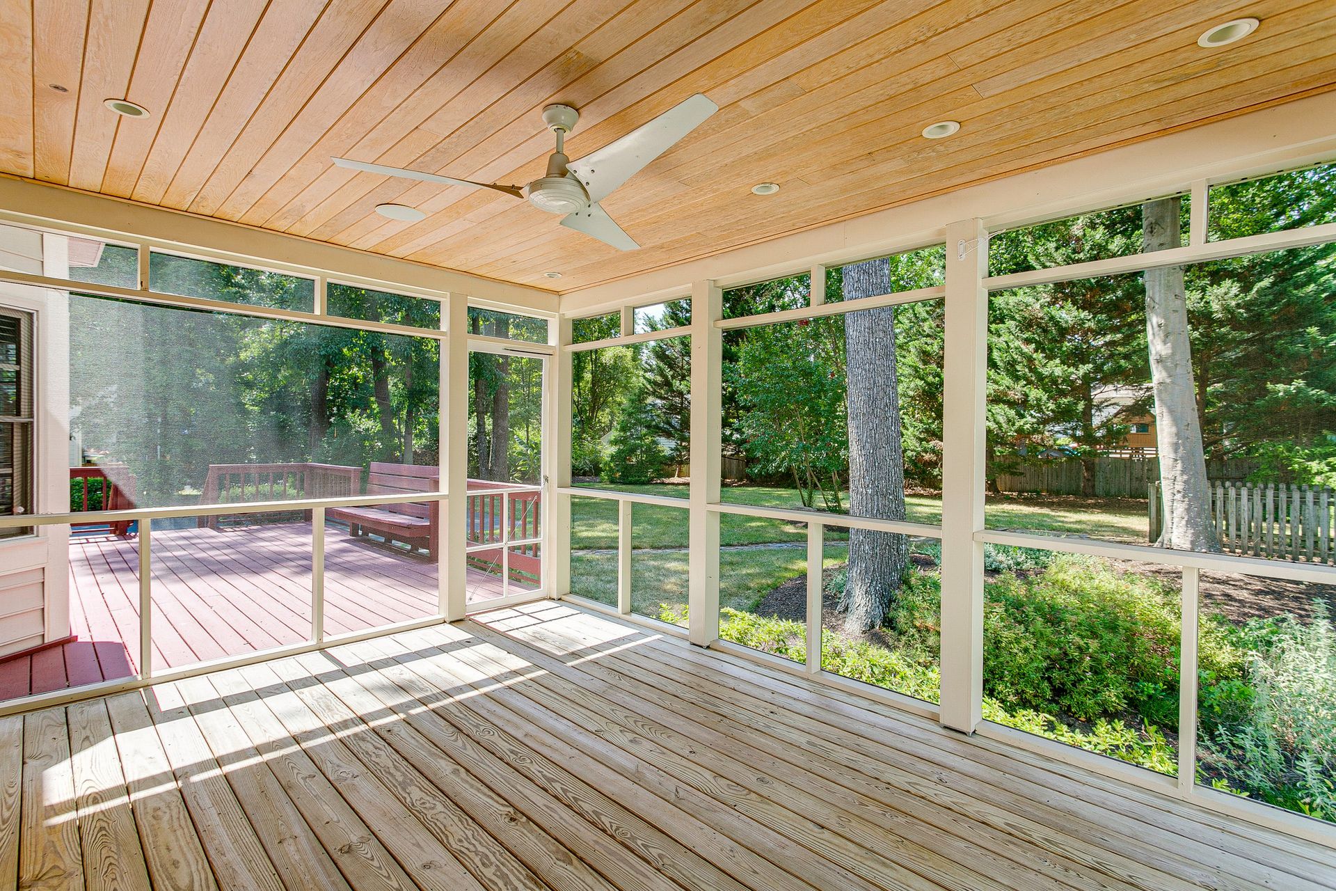 Screened-in porch with wood flooring, ceiling fan, and view of trees and foliage.