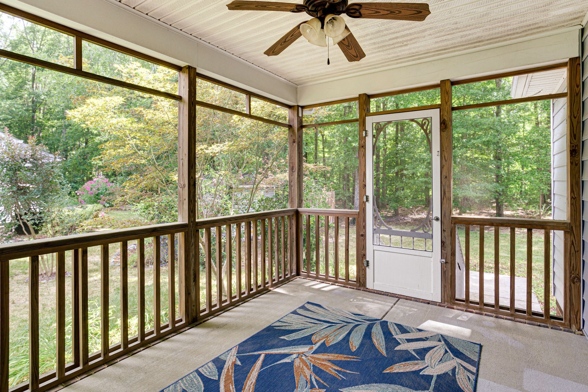 Screened porch with wooden railings, ceiling fan, and blue rug overlooking a wooded yard.