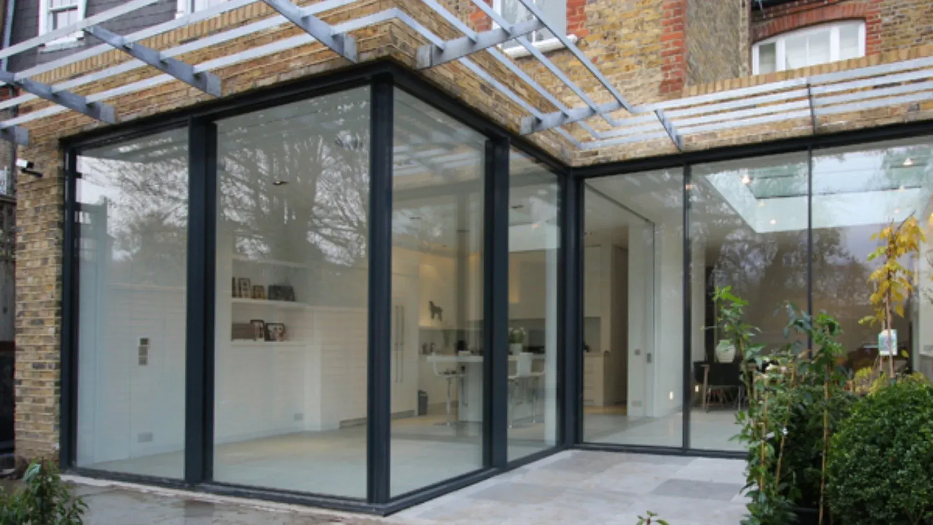 Glass-walled modern home extension with black frames and patio; part of a brick building is visible.