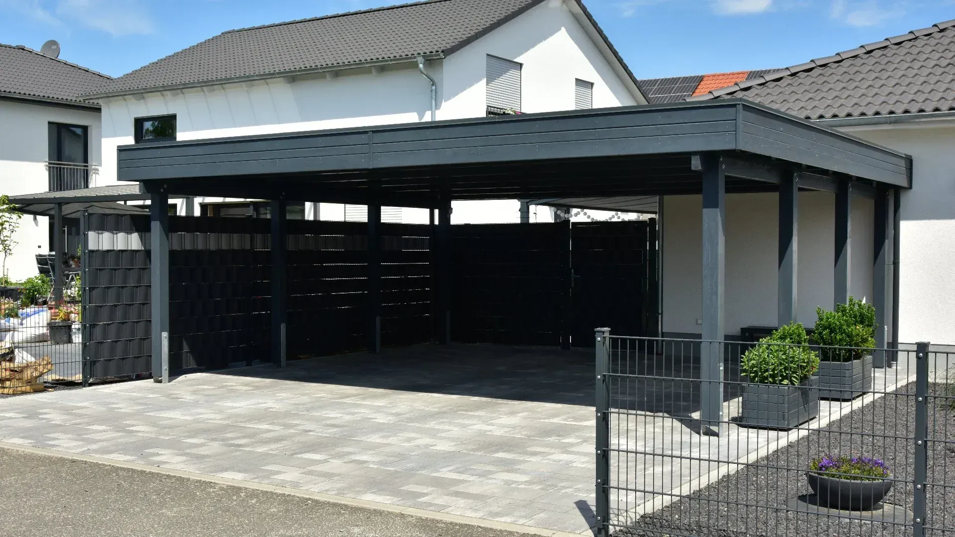 A dark gray carport with a paved floor, black fence and potted plants in front of a house.