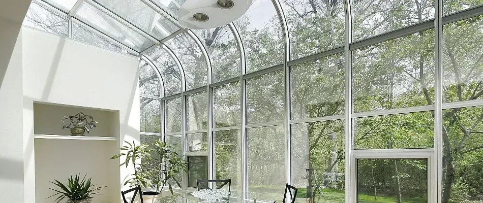 Light-filled sunroom with glass walls and ceiling, looking out onto lush green trees. A table and chairs are inside.