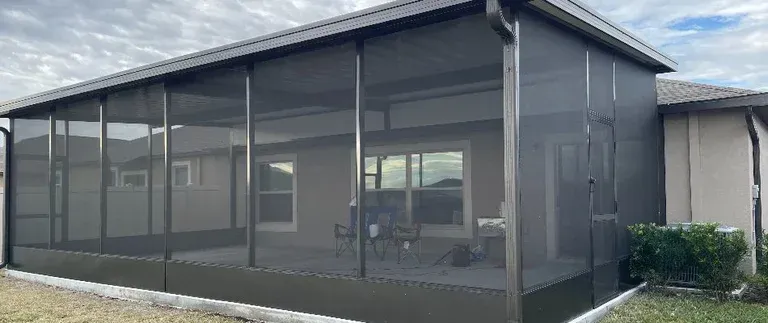 A screened-in porch with dark frames and screens attached to a beige house, cloudy sky in background.