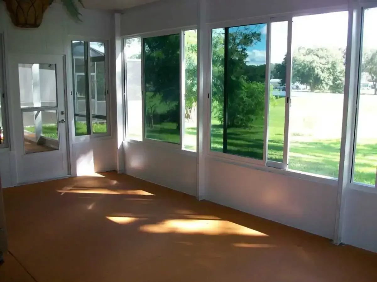 Sunroom interior with tinted windows, overlooking a green lawn. Orange flooring, white walls, and bright sunlight.