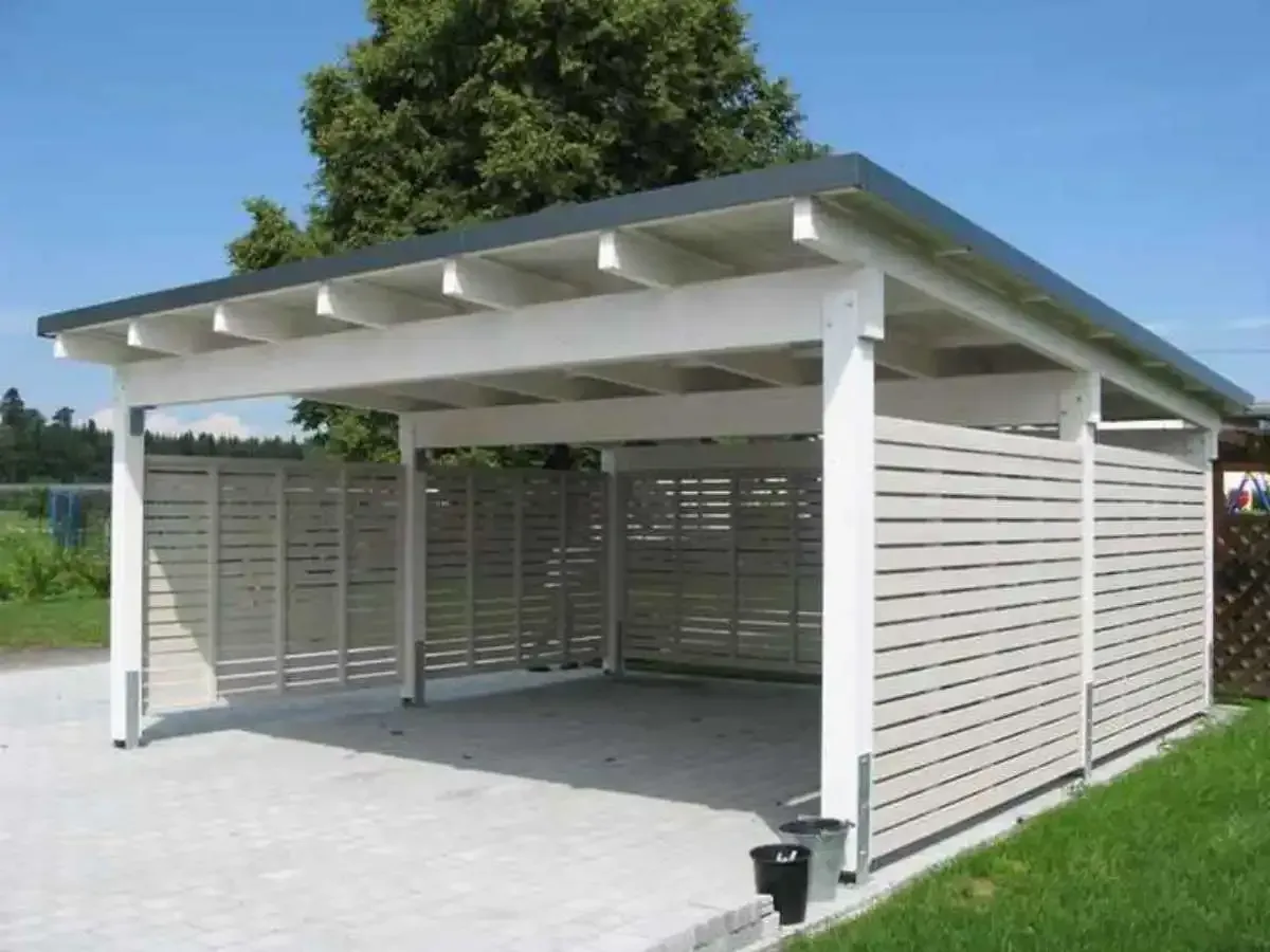 White carport with slatted side walls and a dark roof, built on a concrete slab in a sunny outdoor setting.