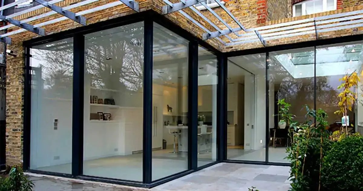 Modern glass-walled extension on brick building. Black frames, light interior, outdoor patio, and metal pergola.