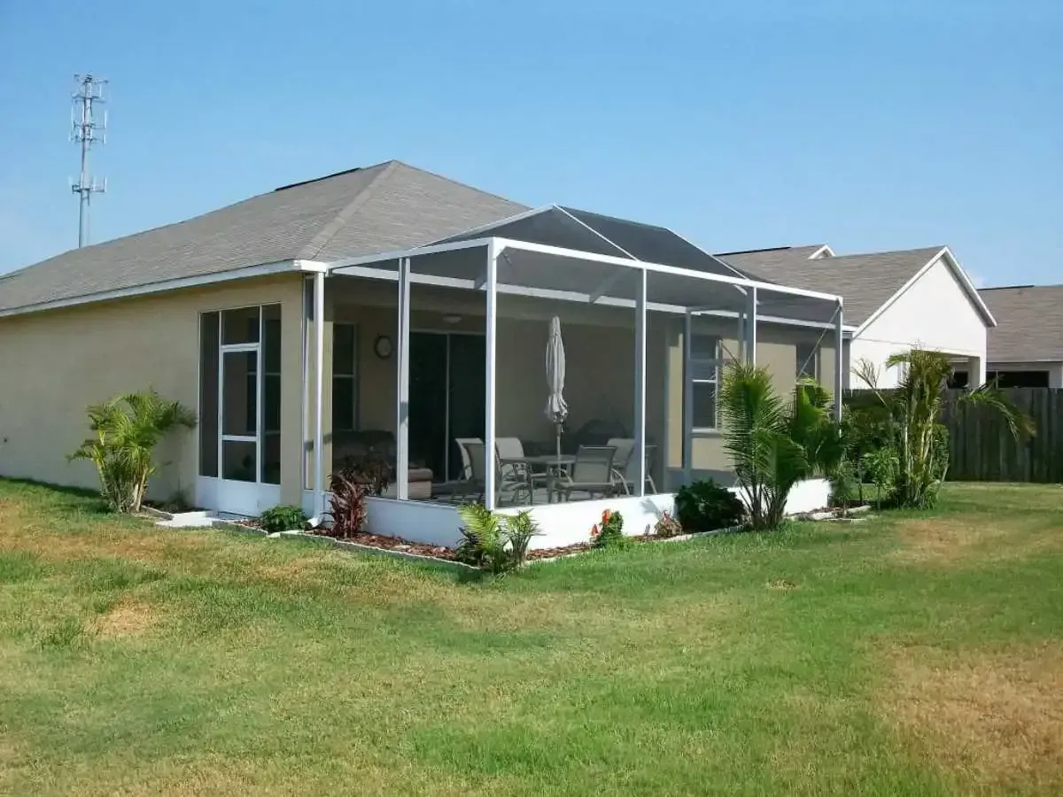 Screened patio attached to a beige house, with outdoor furniture on a grassy lawn under a clear sky.