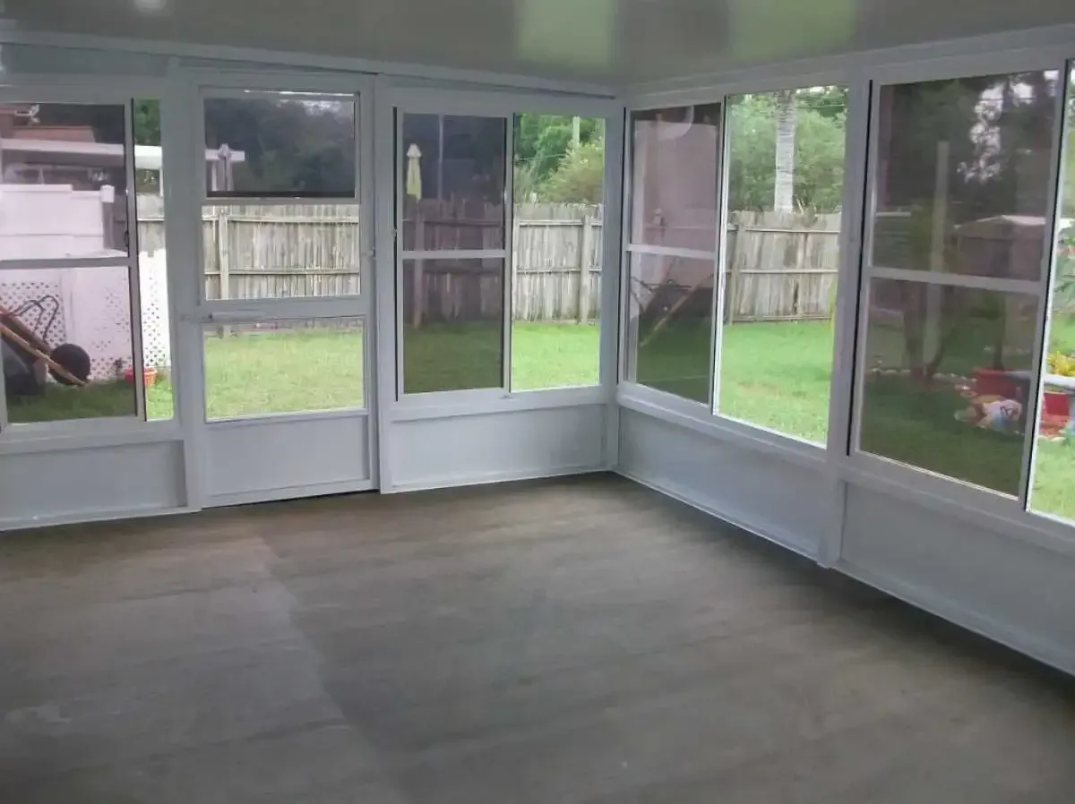 Interior view of a white-framed sunroom with several large windows overlooking a backyard. Empty room with a gray floor.
