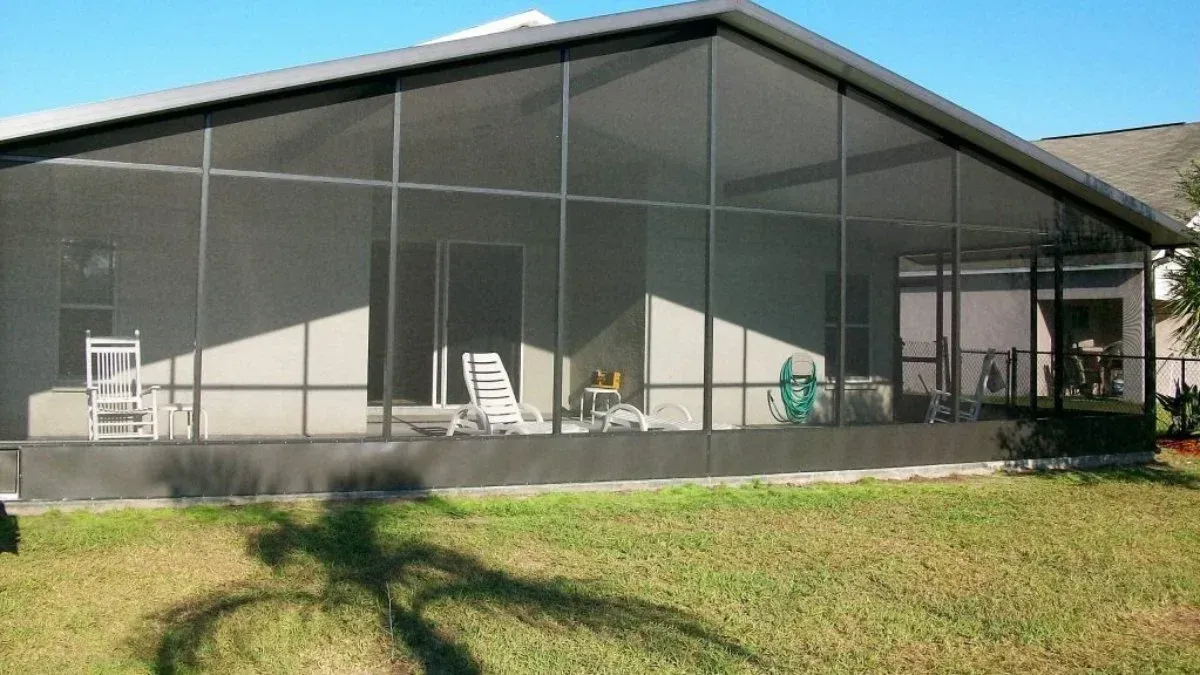 Screened-in patio extension attached to a house. Black framing, beige walls, white chairs, and green grass.