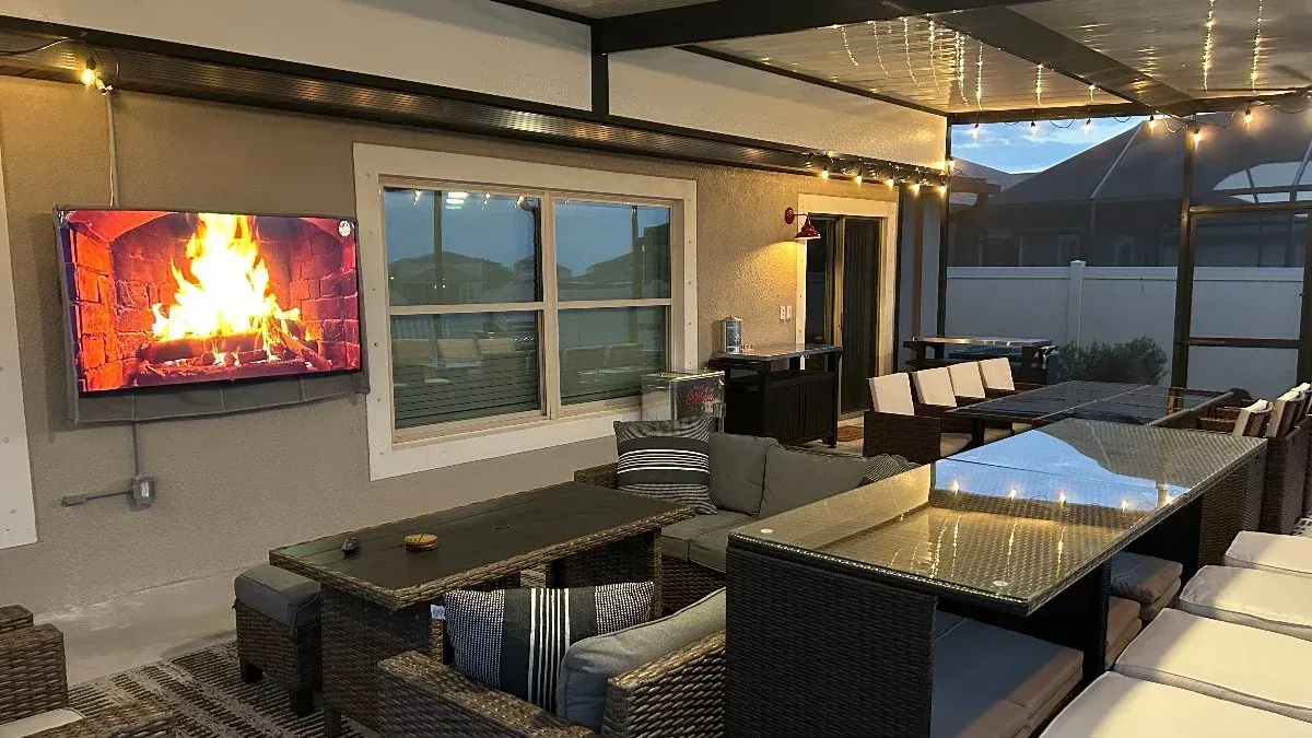 Outdoor patio with a mounted TV showing a fireplace, seating, and string lights.