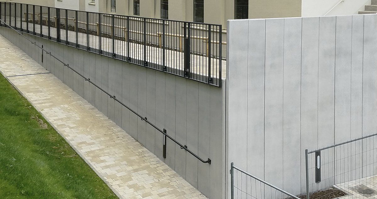 Große L-Betonsteine entlang eines Schulgebäudes. Darauf ist ein schwarzes Geländer montiert