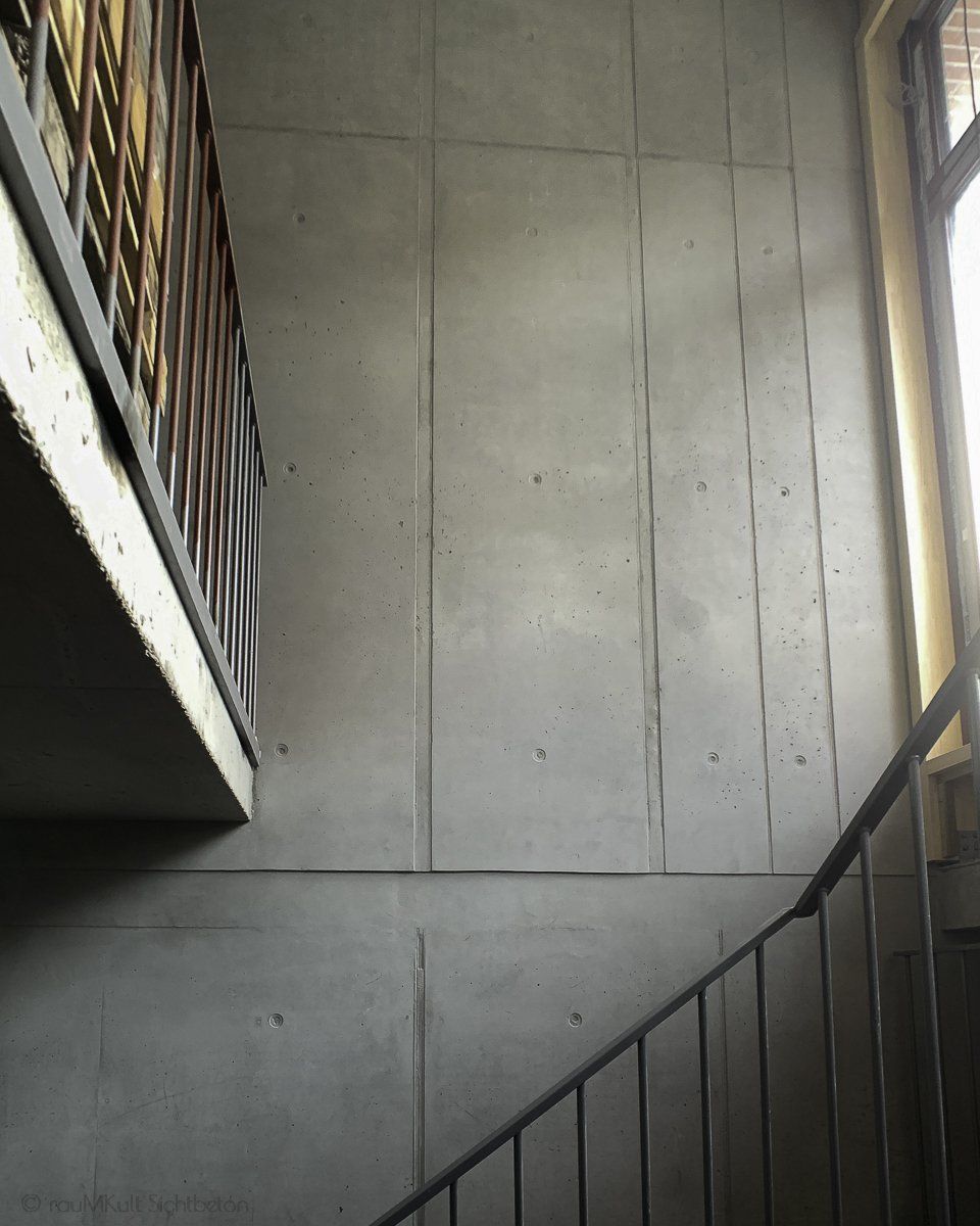 Sichtbeton Kosmetik in einem Treppenhaus von rauMKult Sichtbeton, Hohe Betonwand und ein Geländer in schwarz