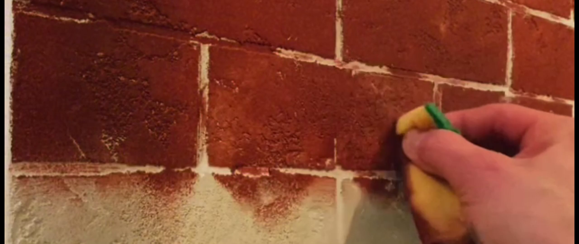 Eine Hand bearbeitet eine Wand mit einem Schwamm, um eine Ziegel-Optik zu erzeugen. Die Wand ist in Rotbrauntönen gestaltet, wobei die Fugenlinien in Weiß die Illusion von Ziegelsteinen verstärken.