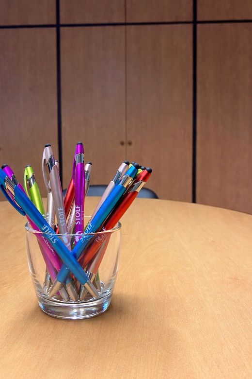 Un bicchiere trasparente pieno di penne colorate di vario tipo, appoggiato su un tavolo di legno in un ufficio.