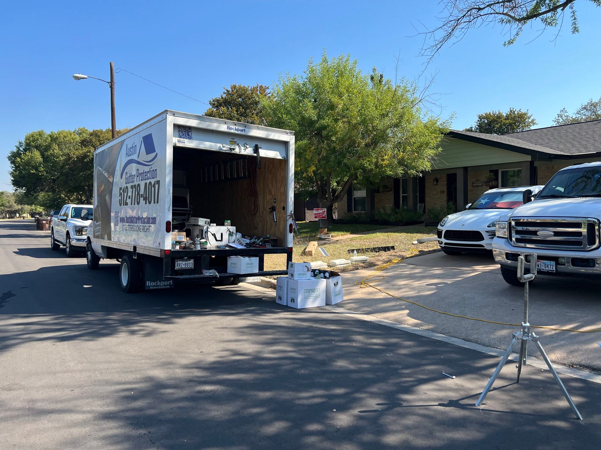 A white moving truck parked on a street in front of a house. Two other vehicles are also parked.