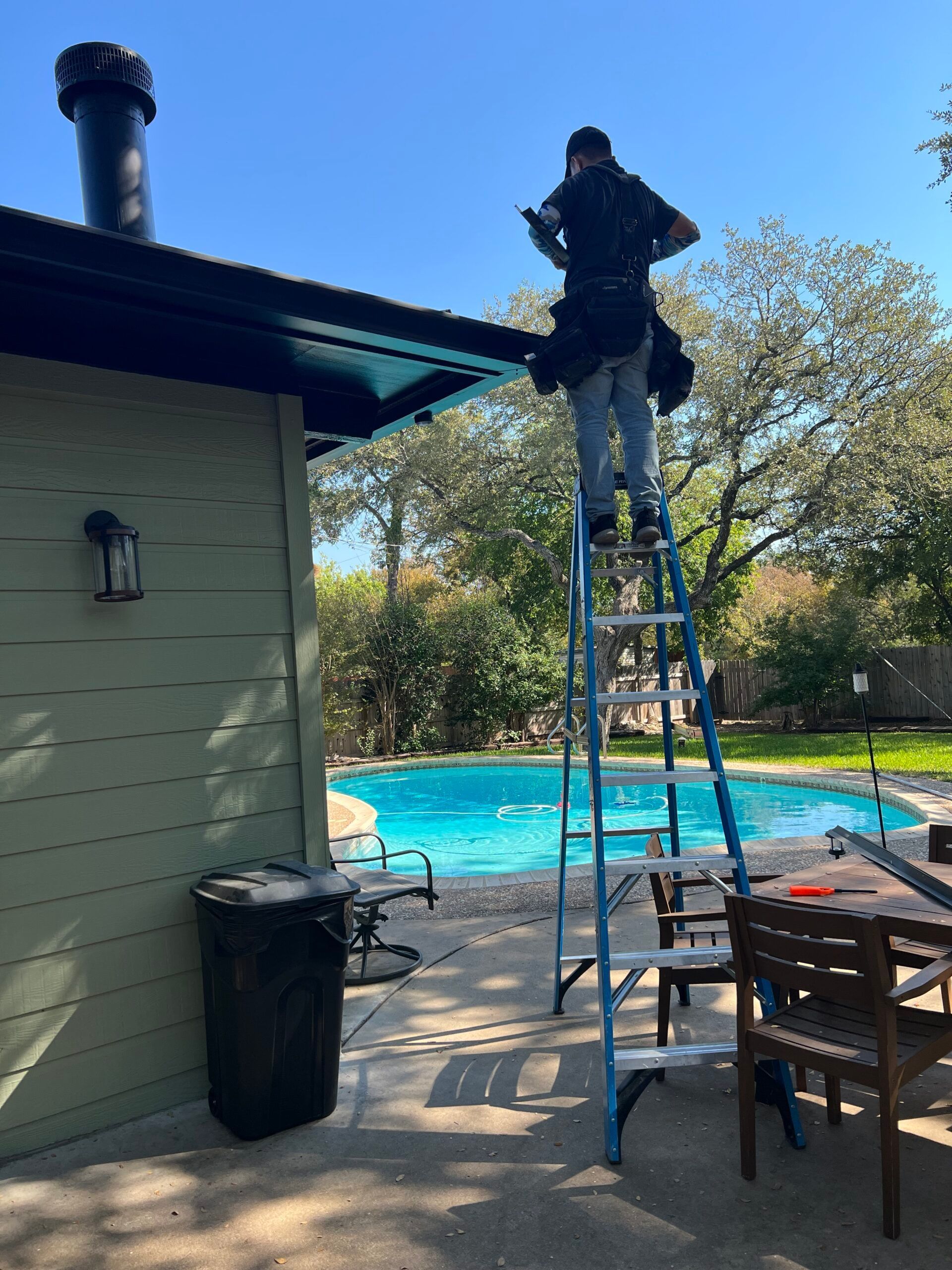 Person on a ladder, cleaning a roof gutter near a pool. Sunny day.
