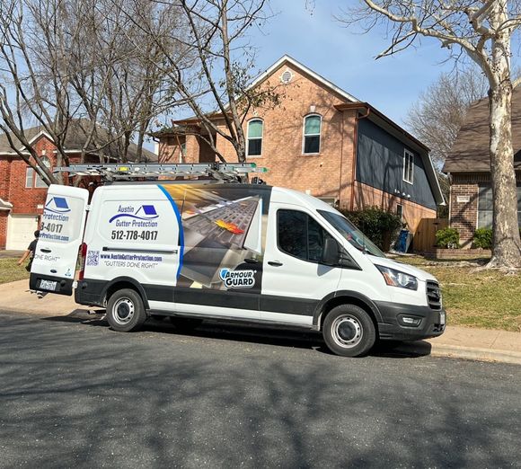White van parked on street with roof company logo, ladder on roof, in front of a house.