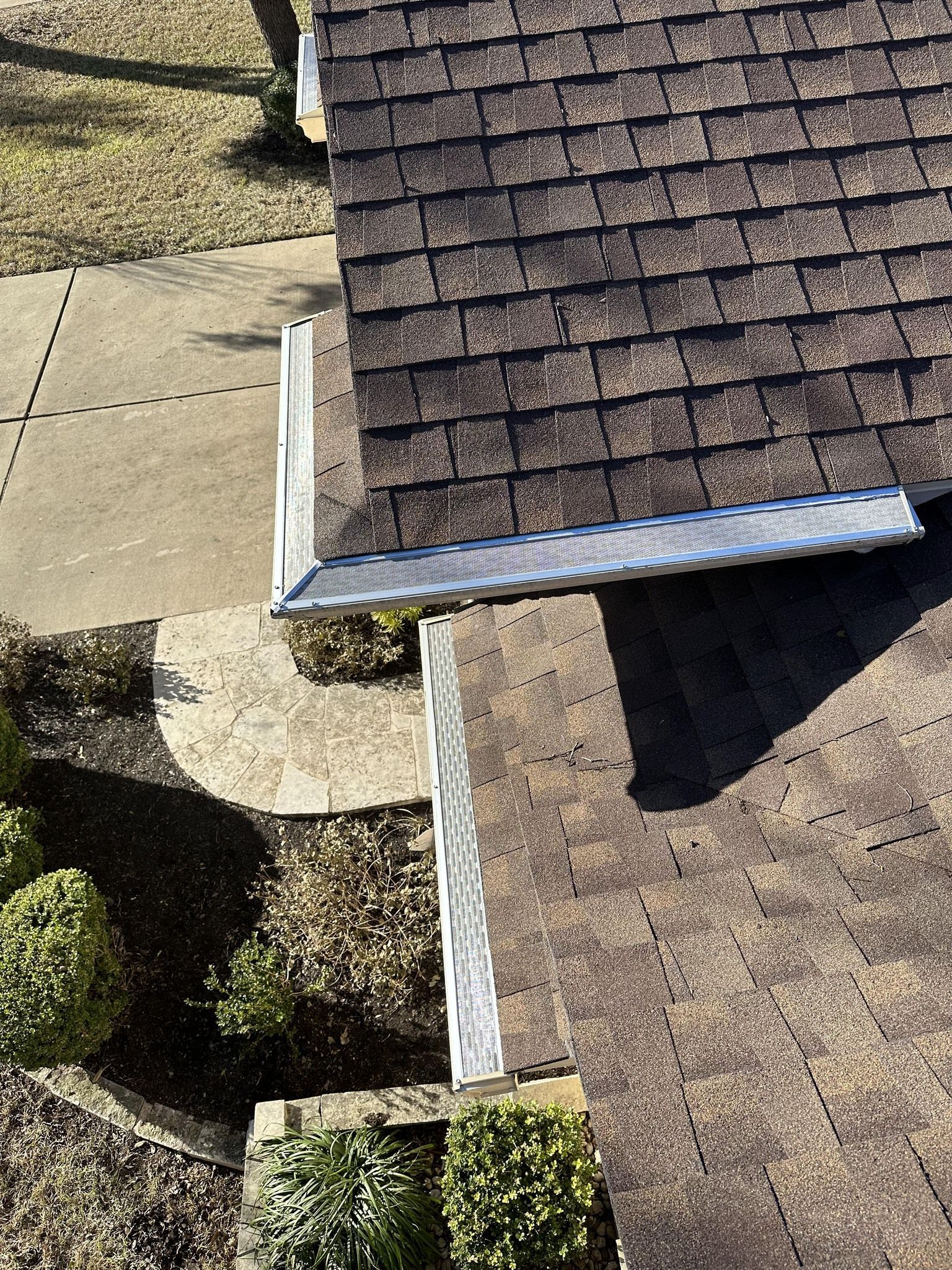 Overhead view of a house roof with dark brown shingles and a silver gutter, next to a concrete sidewalk.