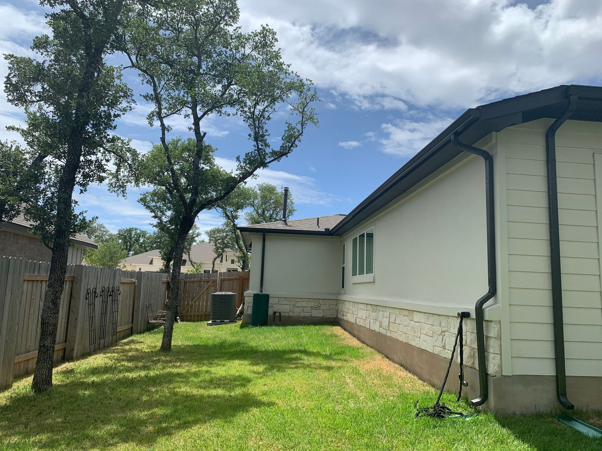 Backyard with trees, green grass, a wooden fence, and a beige house with dark gutters.